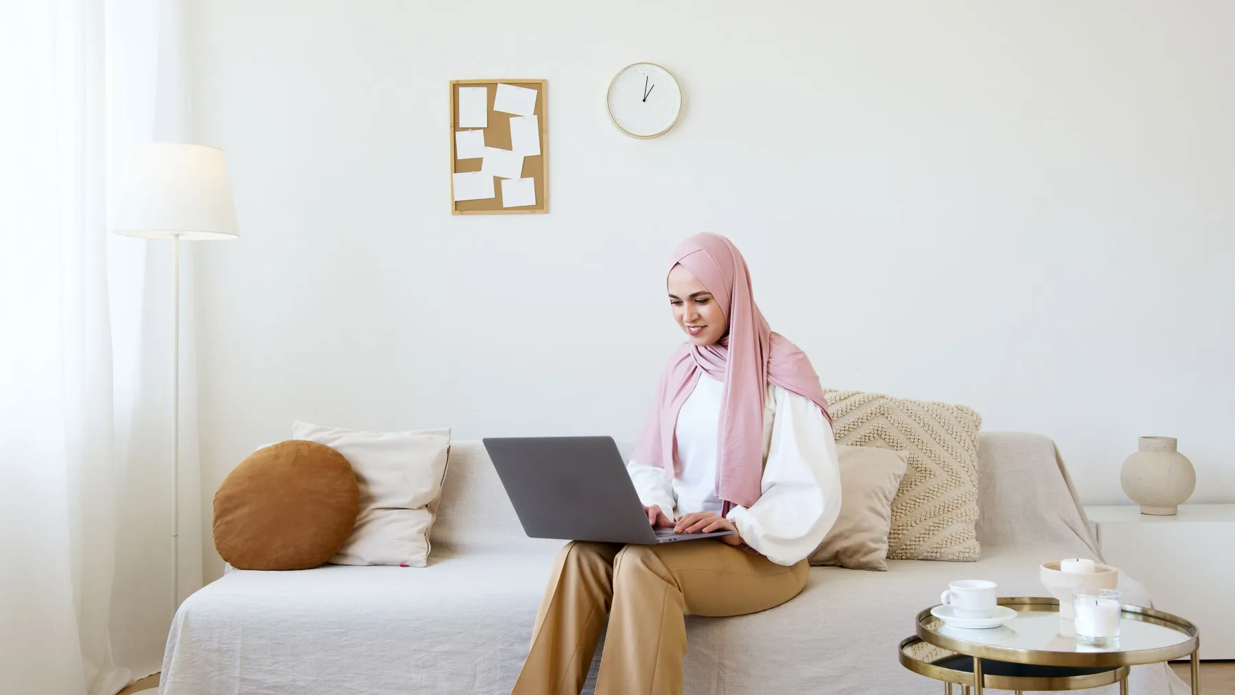 A woman in a pink hijab working on a laptop on a sofa, a cup of coffee on the table beside her