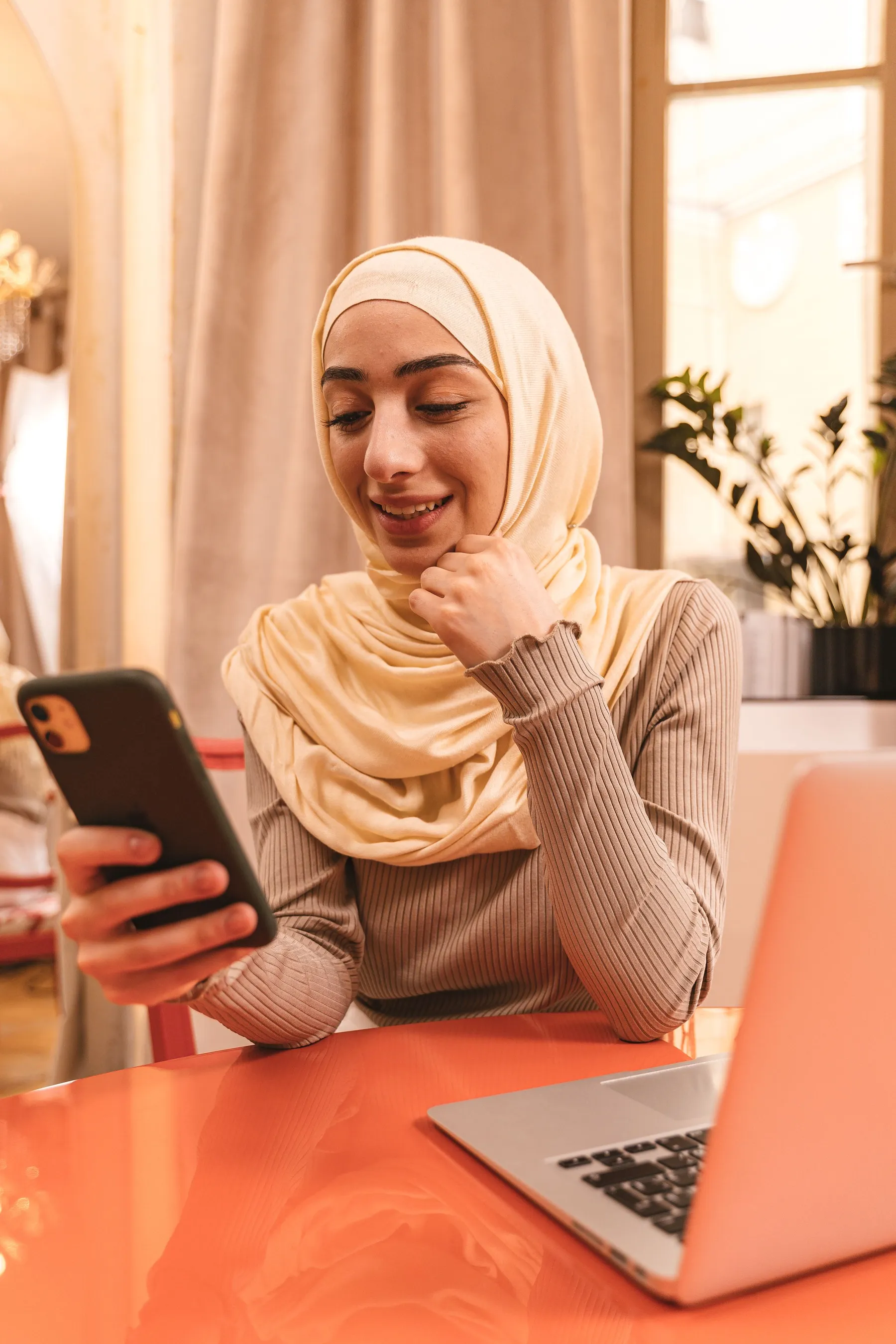 A young woman in a beige hijab smiling at her phone at home