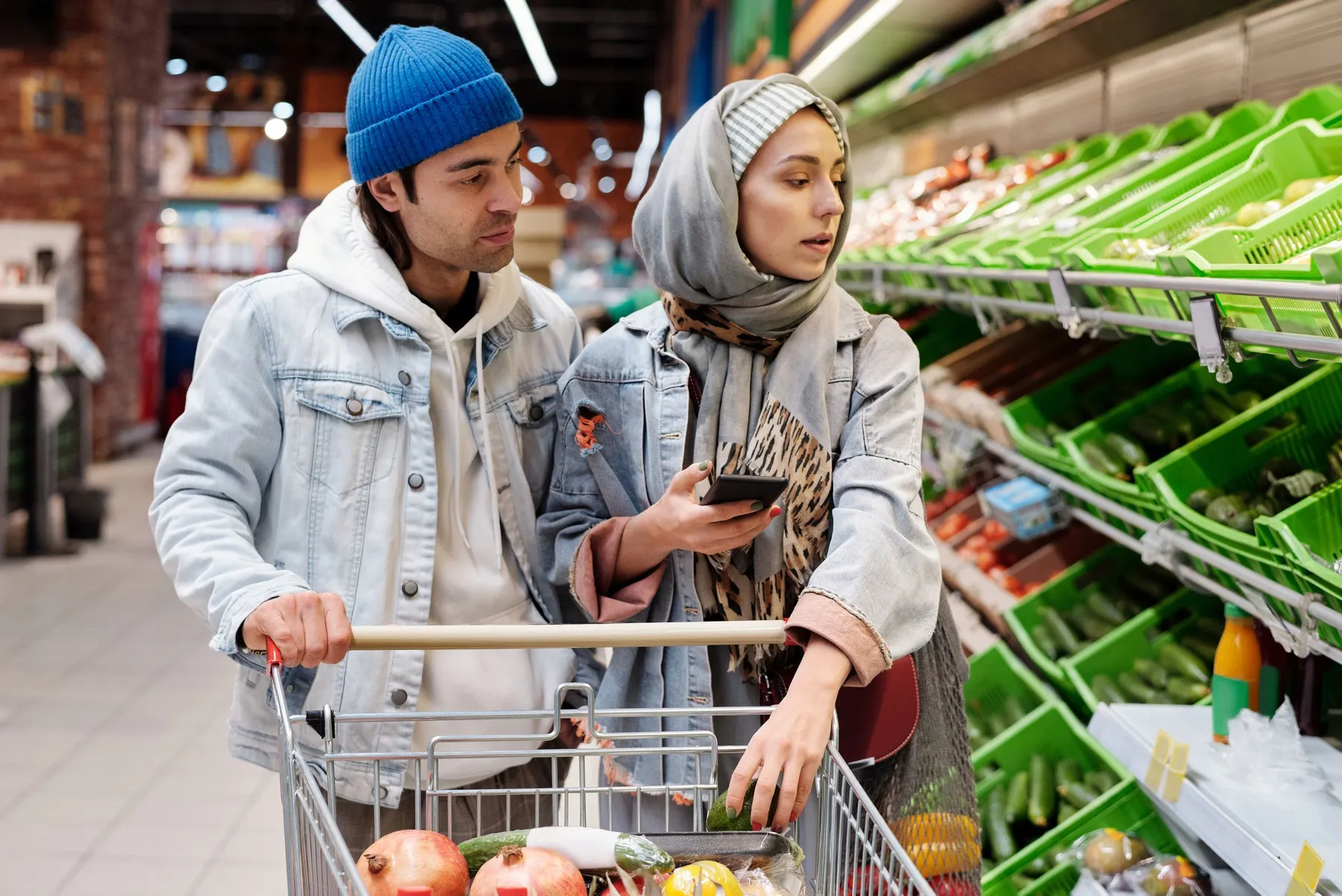 A couple at the supermarket with a trolley; she checks her phone while he leans in to look